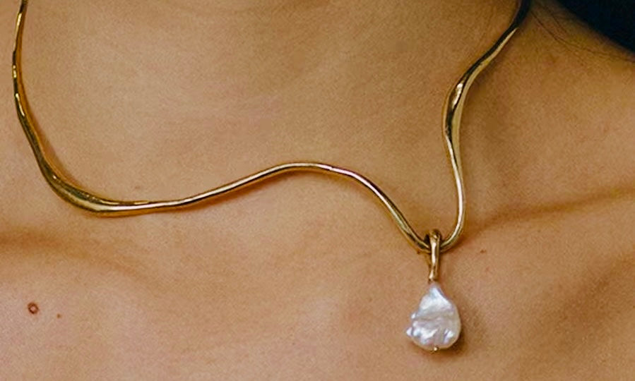 Close-up of a gold pendant necklace featuring an irregular baroque pearl drop on a wavy chain resting against bare skin