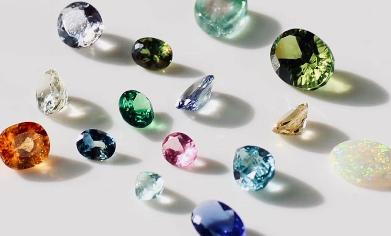 Assorted birthstones in various colours and cuts displayed on a light background, including sapphire, emerald, amethyst, and topaz gemstones