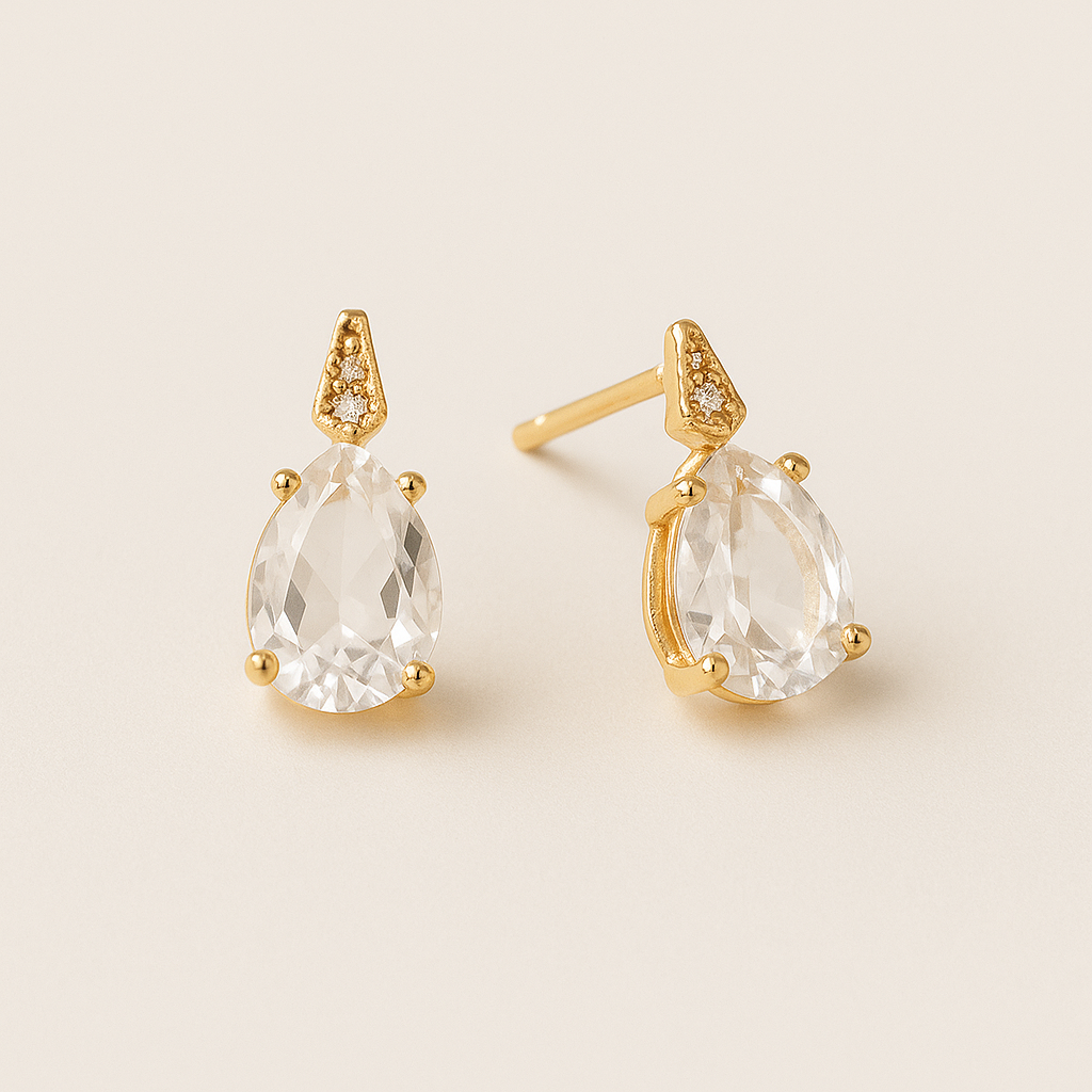 Gold earrings with clear teardrop-shaped gemstones on a beige background the Gembee Jewellery.